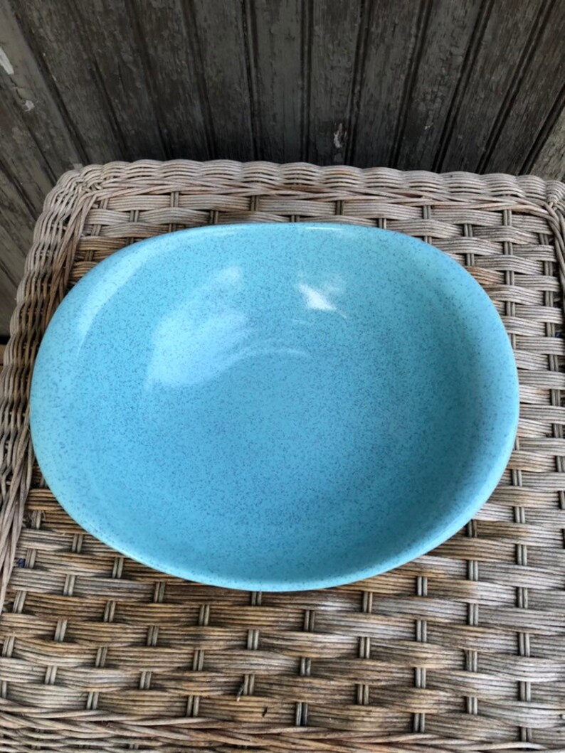 May include: A large, oval-shaped, light blue ceramic bowl with a speckled glaze. The bowl is sitting on a woven wicker surface.