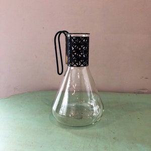 May include: A clear glass pitcher with a black metal handle and a geometric pattern around the top.