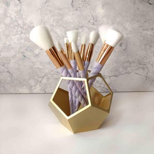 Op de afbeelding: Een gouden geometrische make-up borstelhouder met een set witte en paarse make-up borstels. De borstels hebben roségouden handvatten.