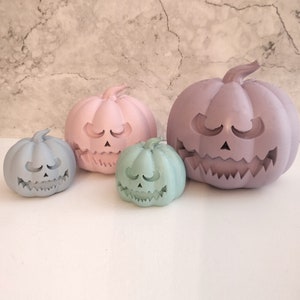 May include: Three pastel-colored ceramic pumpkin decorations. The largest pumpkin is purple, the medium pumpkin is pink, and the smallest pumpkin is light green. All pumpkins have a carved jack-o'-lantern face.