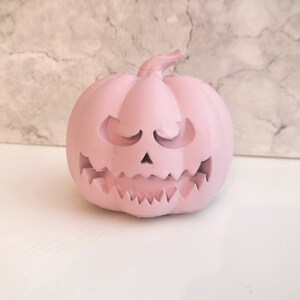 May include: A pink ceramic pumpkin with a carved jack-o'-lantern face. The pumpkin has a wide, toothy grin and closed eyes.