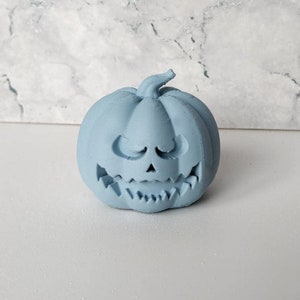 May include: A light blue ceramic pumpkin with a carved jack-o'-lantern face. The pumpkin has a wide, toothy grin and a mischievous expression.