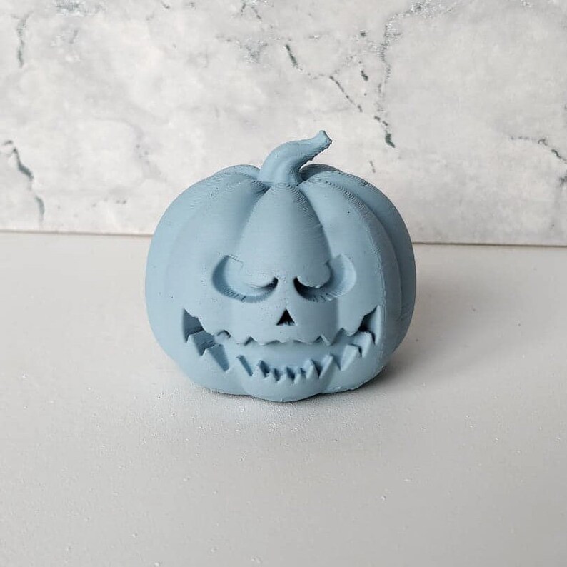 May include: A light blue ceramic pumpkin with a carved jack-o'-lantern face. The pumpkin has a wide, toothy grin and a mischievous expression.