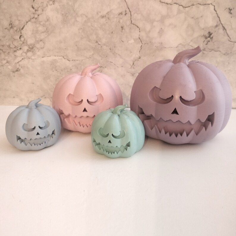 May include: Three pastel-colored ceramic pumpkin decorations. The largest pumpkin is purple, the medium pumpkin is pink, and the smallest pumpkin is light green. All pumpkins have a carved jack-o'-lantern face.