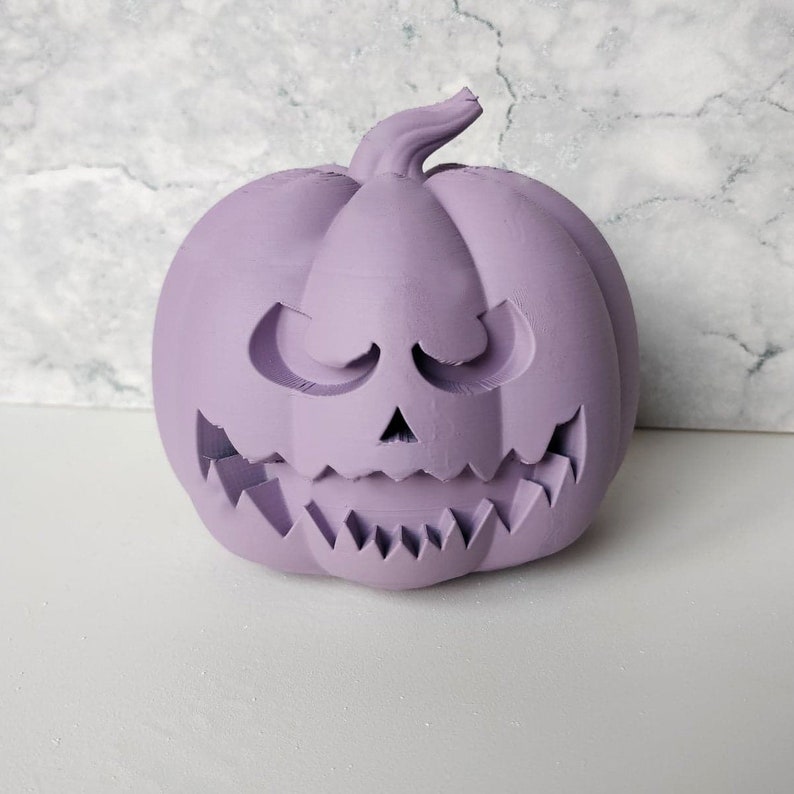 May include: A purple ceramic pumpkin with a carved jack-o'-lantern face. The pumpkin has a wide, toothy grin and a mischievous expression.