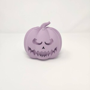 May include: A purple ceramic pumpkin with a carved jack-o'-lantern face. The pumpkin has a wide, toothy grin and small, closed eyes.