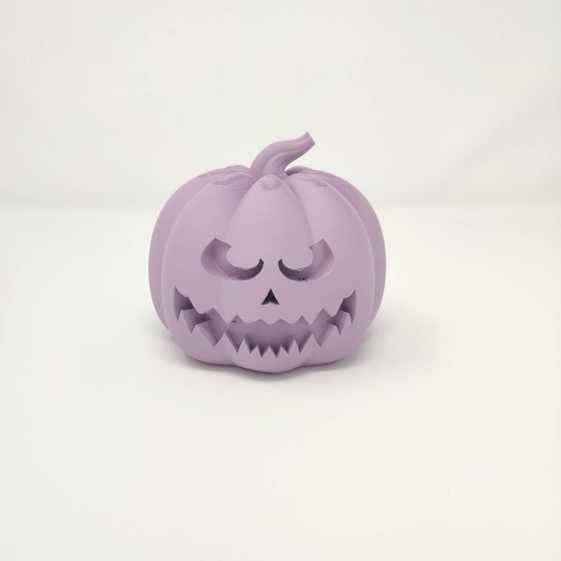 May include: A purple ceramic pumpkin with a carved jack-o'-lantern face. The pumpkin has a wide, toothy grin and small, closed eyes.