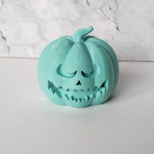 May include: A teal green ceramic pumpkin with a carved jack-o'-lantern face. The pumpkin has a wide, toothy grin and small, closed eyes.