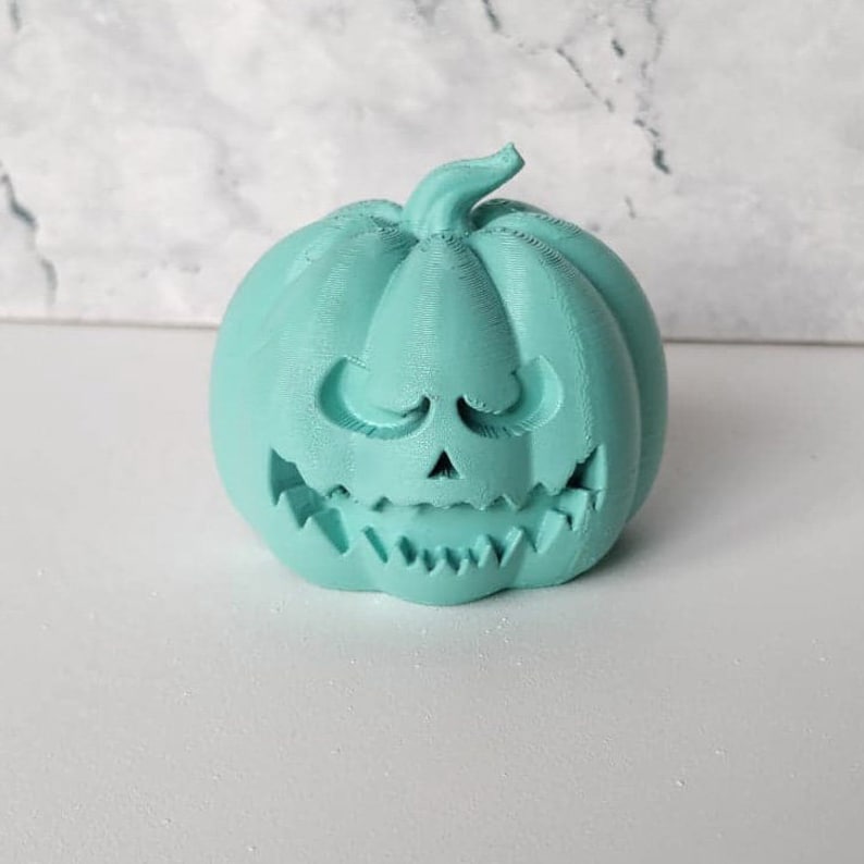 May include: A teal green ceramic pumpkin with a carved jack-o'-lantern face. The pumpkin has a wide, toothy grin and small, closed eyes.