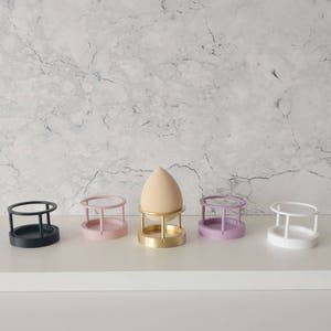 May include: Five makeup sponge holders in different colors: black, pink, gold, purple, and white. The holders are made of plastic and have a circular base with a wire frame.