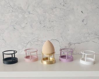 Könnte beinhalten: Fünf Make-up-Schwammbüsten in verschiedenen Farben: Schwarz, Rosa, Gold, Lila und Weiß. Die Halterungen bestehen aus Kunststoff und haben eine runde Basis mit einem Drahtgestell.