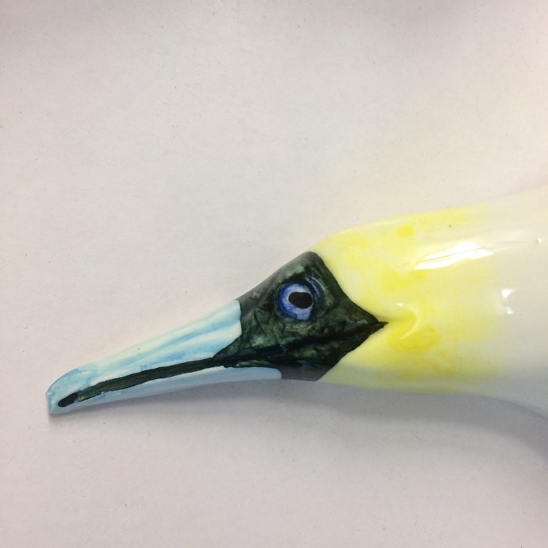 May include: A ceramic bird head with a long, pale blue beak and a yellow and white head. The bird has a dark eye with a blue iris and black markings around the eye.