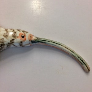 May include: A ceramic bird sculpture with a long, curved beak. The bird is painted in shades of brown, white, and green, with an orange eye.