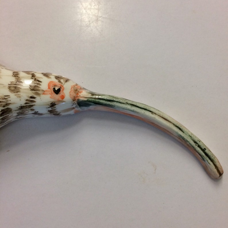 May include: A ceramic bird sculpture with a long, curved beak. The bird is painted in shades of brown, white, and green, with an orange eye.