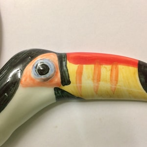 May include: Close-up of a ceramic toucan figurine with a black, white and yellow beak. The toucan's eye is painted blue with a black pupil.