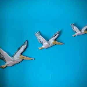 May include: Three ceramic pelican wall sculptures in flight against a bright blue background. The pelicans are painted white with black and grey markings and yellow beaks.