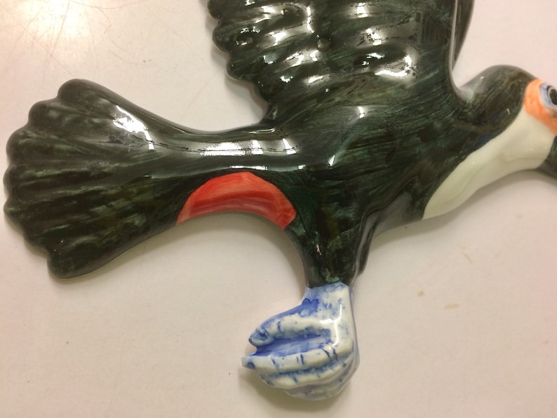 May include: A ceramic toucan figurine painted in black, green, white, red, and blue. The toucan is in flight with its wings outstretched. The toucan's beak is open and its eyes are closed.