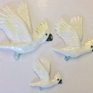 May include: Three white ceramic cockatoo bird figurines in flight. The largest cockatoo is in the centre of the image, with a smaller cockatoo to the right and the smallest cockatoo to the left. The cockatoos have yellow accents on their wings and heads.