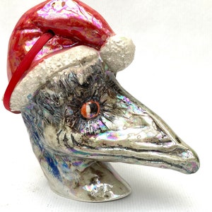 Puede incluir: Una escultura de cerámica de la cabeza de un pájaro que lleva un gorro de Papá Noel rojo con ribete blanco. La cabeza del pájaro es iridiscente y tiene un pico largo y puntiagudo.