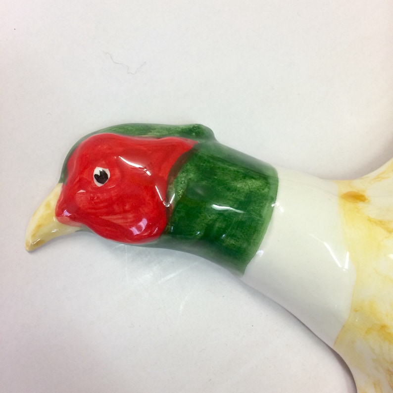 May include: A ceramic pheasant figurine, featuring a red head, green neck, and white body. The beak is yellow. The bird's head is turned to the side, showcasing its detailed features. The figurine is likely a decorative item.