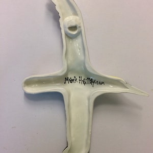 May include: A ceramic bird sculpture in flight, viewed from the back. The sculpture is mainly off-white with black wingtip details. The text "Mark Hailley.com" is written in black script. A hole is present for hanging.