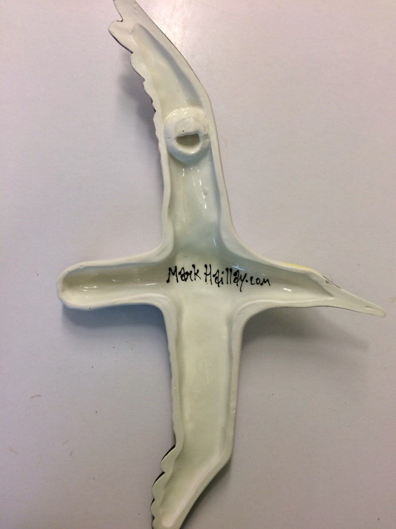 May include: A ceramic bird sculpture in flight, viewed from the back. The sculpture is mainly off-white with black wingtip details. The text "Mark Hailley.com" is written in black script. A hole is present for hanging.