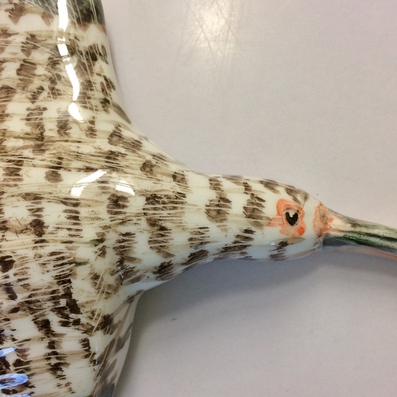 May include: A ceramic sculpture of a bird with brown and white patterned feathers and an orange eye. The bird's beak is long and thin, and it is painted in a realistic style.
