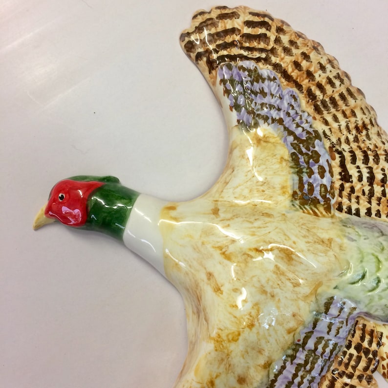 May include: A ceramic pheasant figurine in flight, featuring a red head, green neck, and cream-colored body. The wings display brown and purple feather details. The bird is a decorative item.