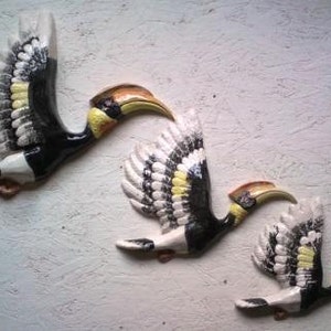 May include: Three ceramic wall sculptures of hornbills in flight. The birds are painted in black, white and yellow with detailed feather patterns.
