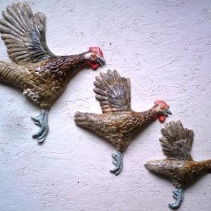 May include: Three ceramic wall sculptures of brown and white chickens with red combs. The chickens are in flight with their wings spread out.
