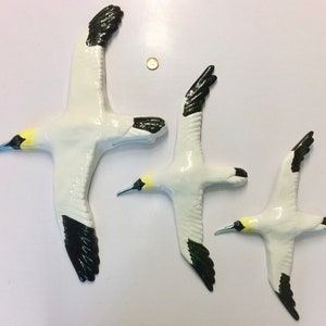 May include: Three ceramic bird sculptures in flight. The birds are white with black wingtips and tail feathers. They have yellow and black beaks. The sculptures vary in size, with the largest bird measuring approximately 30 cm in wingspan.