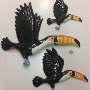May include: Three ceramic toucans in flight, painted in black, white, orange, and yellow. The toucans are in different sizes and are arranged in a staggered formation.