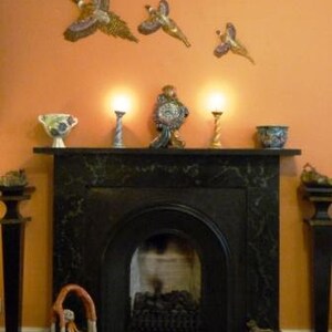 May include: A black marble fireplace with a decorative mantel, featuring a clock, vases, and candle holders. Above, three decorative bird sculptures are mounted on an orange wall. A sculpture of a person bending over is in front of the fireplace.