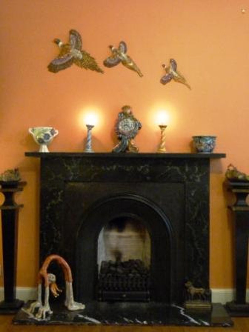 May include: A black marble fireplace with a decorative mantel, featuring a clock, vases, and candle holders. Above, three decorative bird sculptures are mounted on an orange wall. A sculpture of a person bending over is in front of the fireplace.