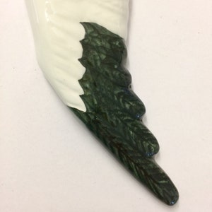 May include: A ceramic wing-shaped ornament. The wing is primarily white with a glossy finish, and a section of dark green, leaf-like detailing. The ornament has a smooth, curved shape and is likely for decorative purposes.