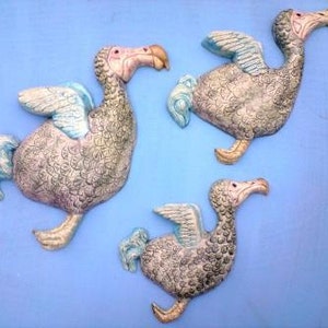 May include: Three ceramic wall sculptures of extinct dodo birds with blue and grey wings. The birds are in flight with their beaks open.