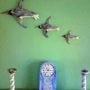 May include: Three ceramic penguin figurines in flight, painted in shades of grey and black, with yellow accents. The penguins are mounted on a green wall, with a blue clock and two decorative candle holders on a wooden shelf below.