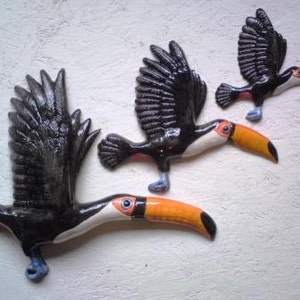 May include: Three ceramic toucan bird sculptures. The toucans are black with white and orange beaks. The toucans are in flight with their wings spread out.