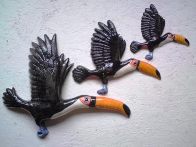 May include: Three ceramic toucan bird sculptures. The toucans are black with white and orange beaks. The toucans are in flight with their wings spread out.