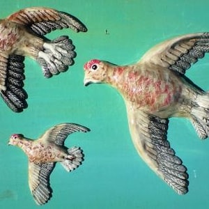 May include: Three ceramic sculptures of birds in flight. The birds are painted in shades of brown, white, and pink. The birds are all facing the same direction and have their wings spread out.
