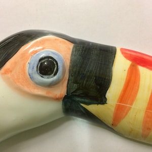 May include: Close-up of a ceramic toucan sculpture with a black, orange, and yellow beak. The toucan's eye is painted blue with a black pupil.
