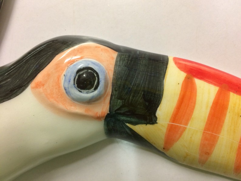 May include: Close-up of a ceramic toucan sculpture with a black, orange, and yellow beak. The toucan's eye is painted blue with a black pupil.