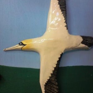 May include: A ceramic sculpture of a bird in flight, with a white body, black wingtips, and a yellow head. The bird has a long beak and is set against a blue and green backdrop, evoking a sky and landscape scene.