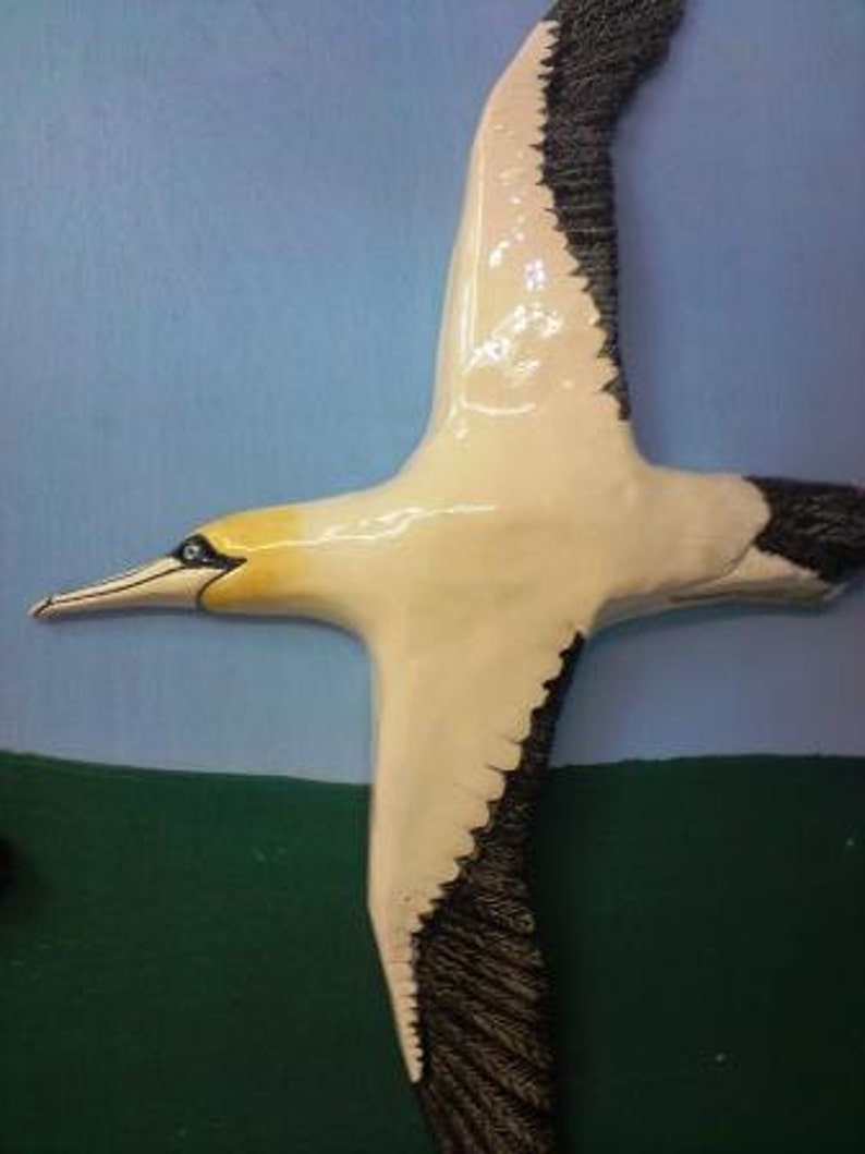 May include: A ceramic sculpture of a bird in flight, with a white body, black wingtips, and a yellow head. The bird has a long beak and is set against a blue and green backdrop, evoking a sky and landscape scene.