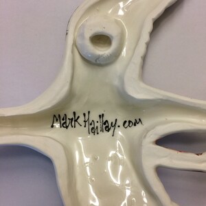 May include: White ceramic wall hanging in the shape of a bird with a hole in the back for hanging. The bird has a black signature that reads "Mark Haillay.com".