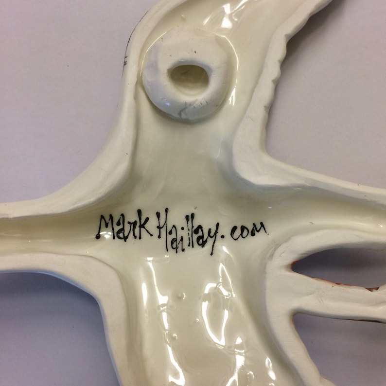 May include: White ceramic wall hanging in the shape of a bird with a hole in the back for hanging. The bird has a black signature that reads "Mark Haillay.com".