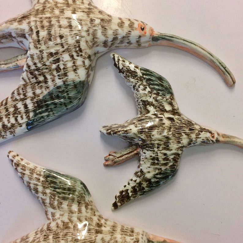 May include: Three ceramic sculptures of brown and white birds with long beaks and green accents. The birds are in flight and appear to be made of a textured material.