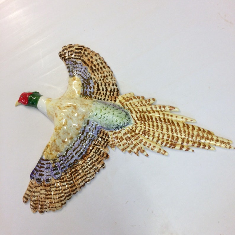 May include: A ceramic pheasant in flight, featuring a detailed design. The bird has brown, tan, and purple wings, a cream-colored body, and a red and green head. The pheasant is a decorative item.