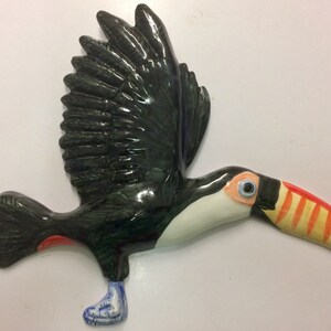 May include: A ceramic toucan figurine painted in black, green, white, orange, and red. The toucan is in flight with its wings spread and its beak open.