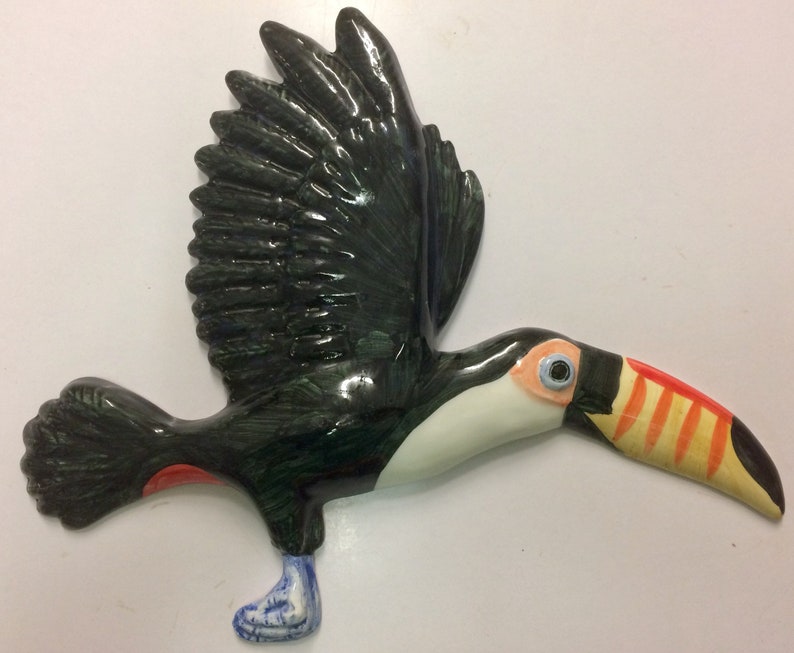 May include: A ceramic toucan figurine painted in black, green, white, orange, and red. The toucan is in flight with its wings spread and its beak open.